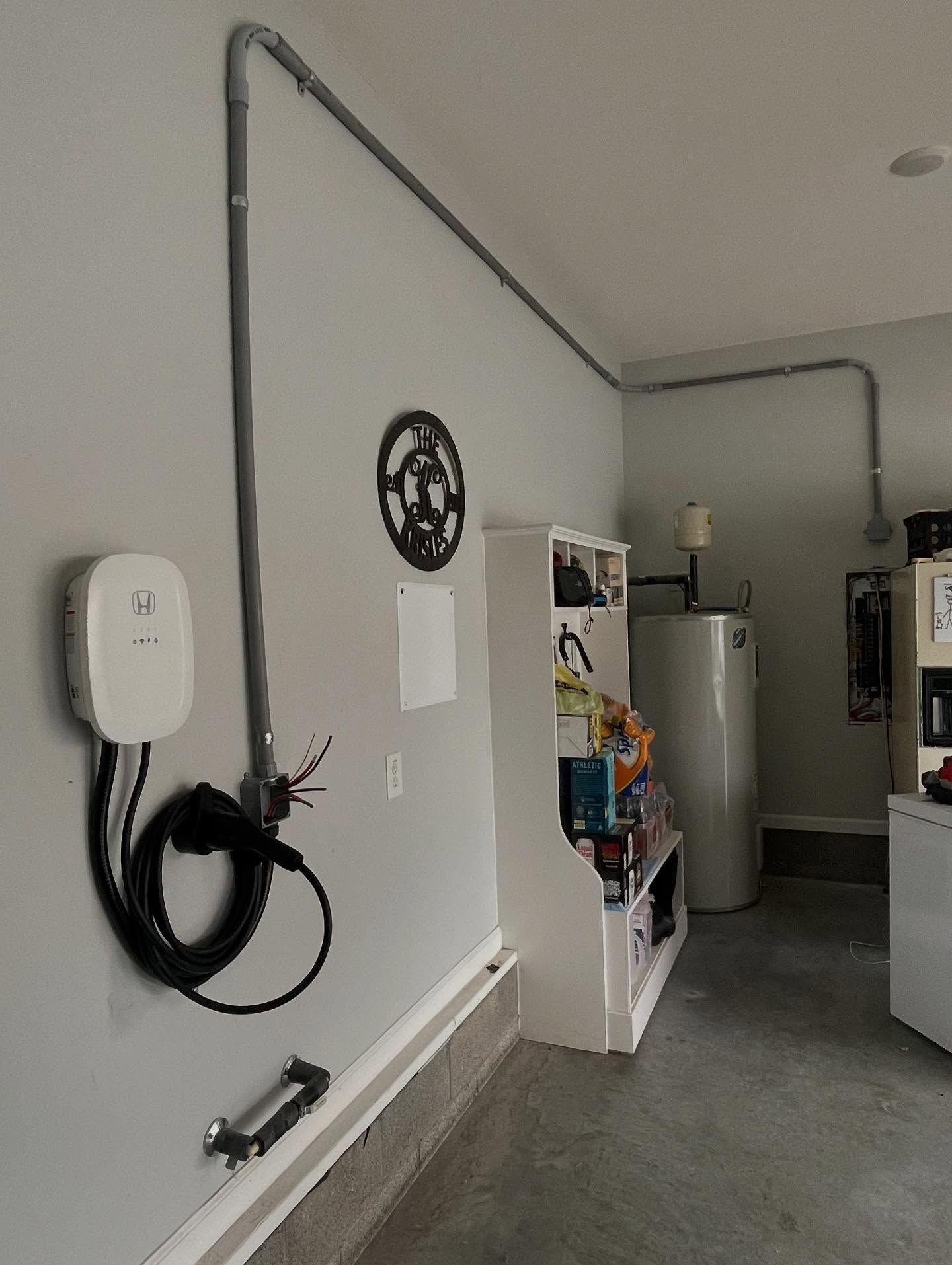 White EV charger on a gray wall with surface conduit running overhead in a utility room.