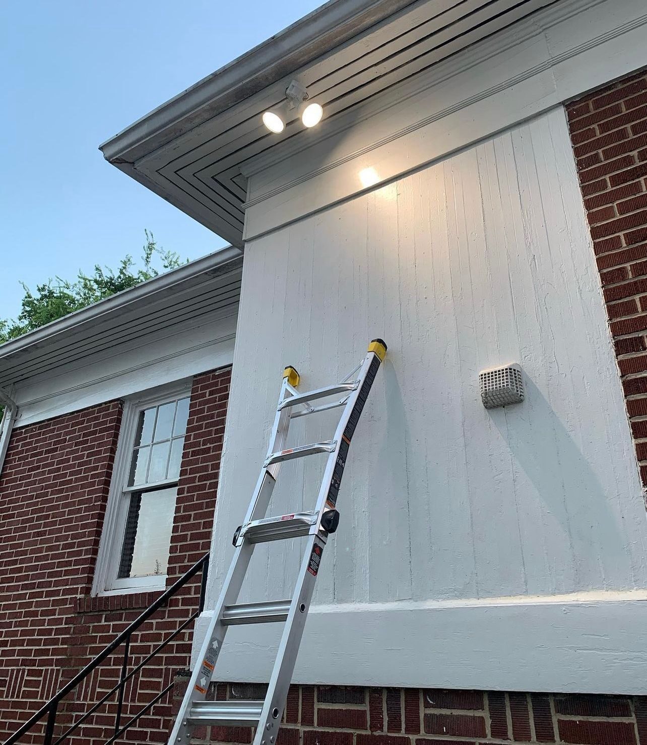Two floodlights mounted under the eave of a brick and white-paneled building with a ladder beside the wall