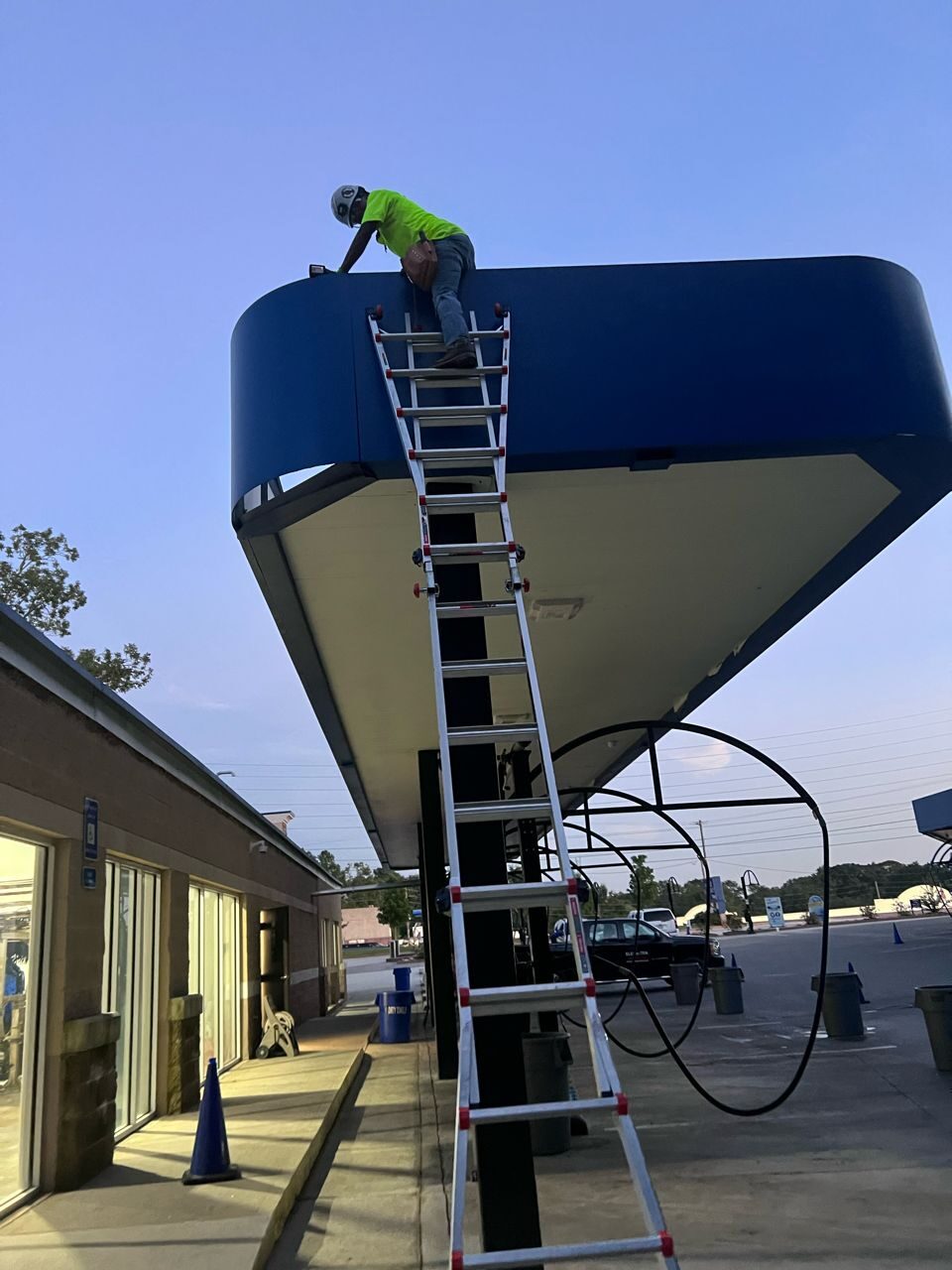 Technician on a long ladder at the edge of a large blue-trimmed canopy with hoses hanging below