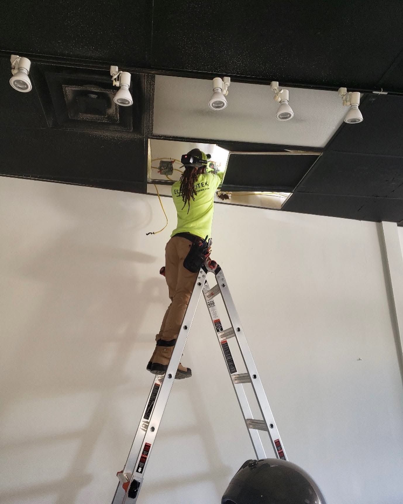 Technician on a stepladder working in a ceiling opening beside a row of track light heads on a black ceiling