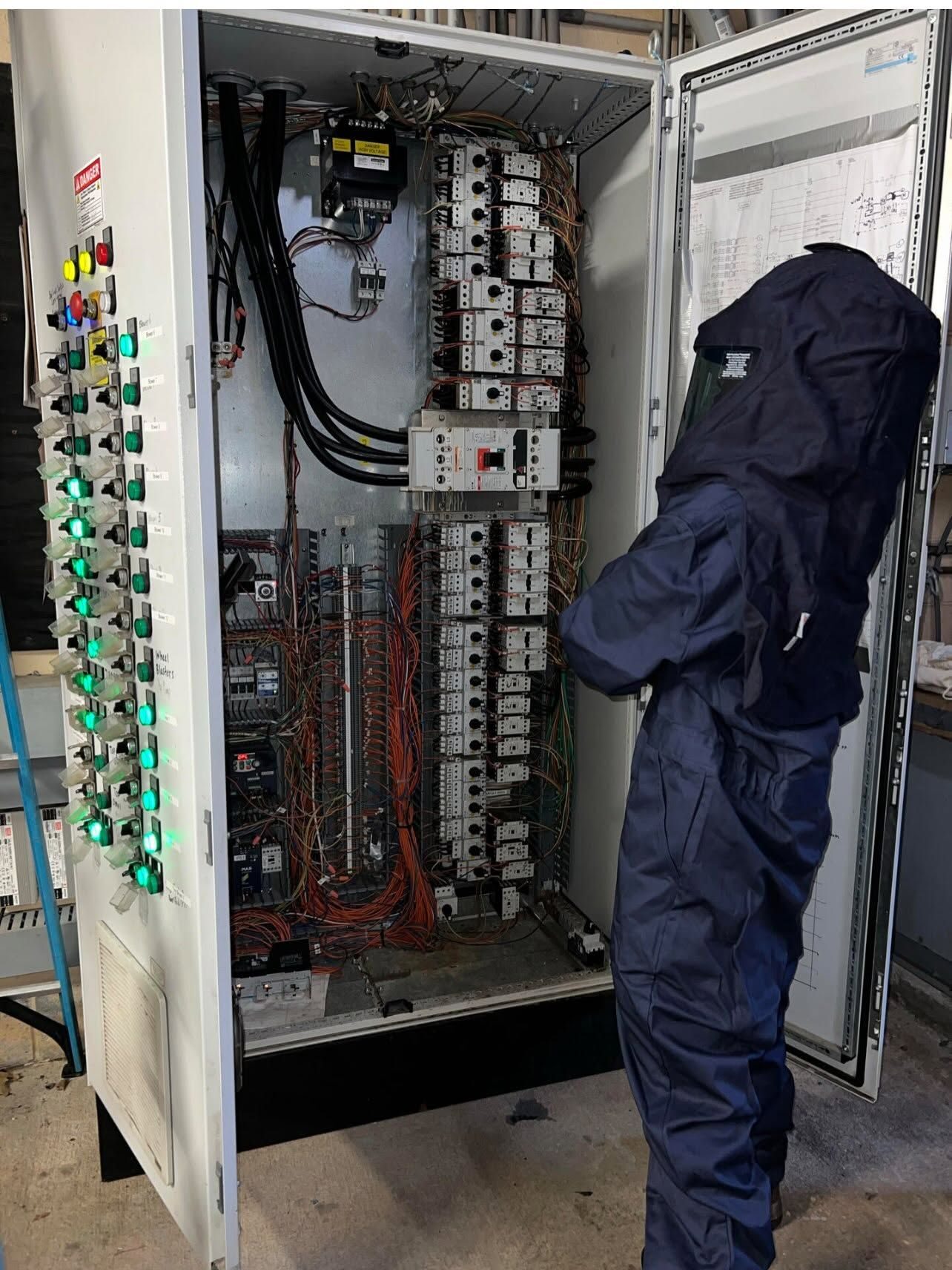 Technician in a full arc-rated suit and hood working at an open motor control cabinet with rows of green pilot lights and dense wiring