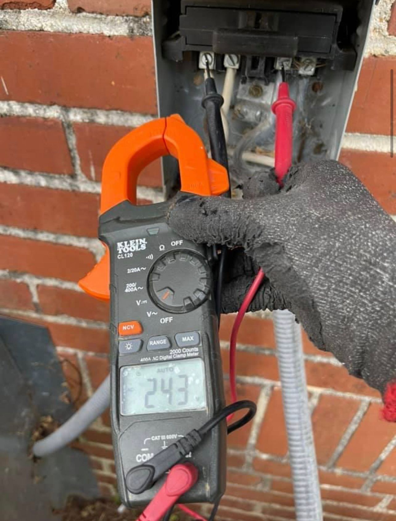 Gloved hand holding a clamp meter reading 243 volts at an open meter base on a brick wall.