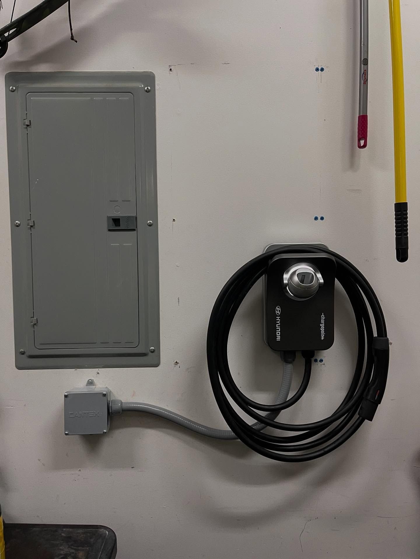 Wall-mounted EV charger with coiled cable below a gray breaker panel, with conduit running between them