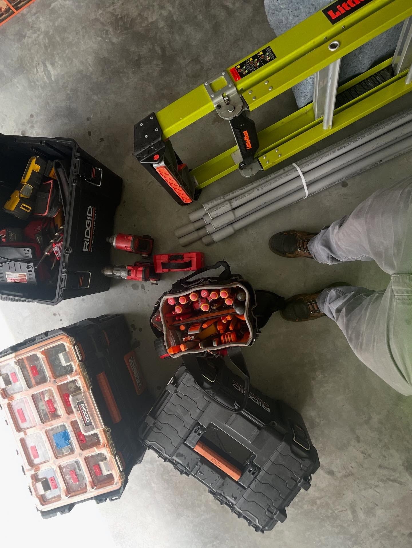 Overhead view of a fiberglass ladder, tool cases, an open tool bag, and lengths of conduit on a concrete floor.