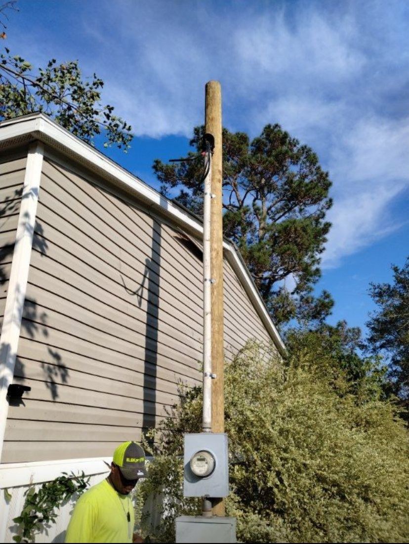 Wooden service pole with a meter base beside the side of a manufactured home under a blue sky.