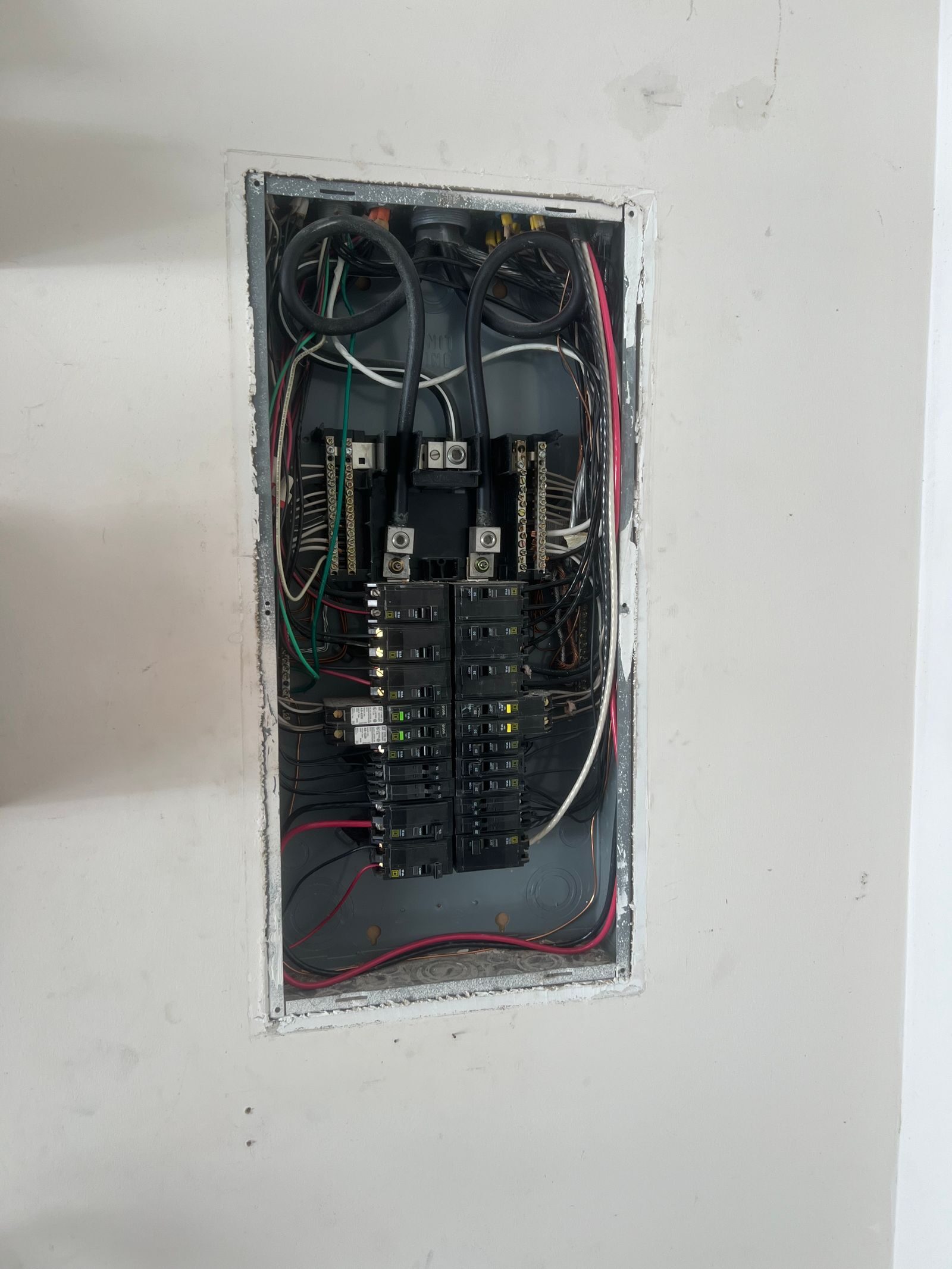 Recessed breaker panel open in a white wall, showing breakers, bus bars, and incoming cables.