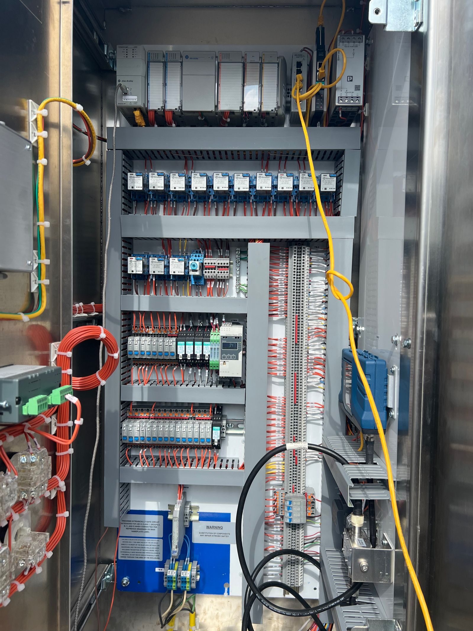 Stainless steel control cabinet opened to show PLC modules, relay banks and terminal blocks with a yellow patch cable