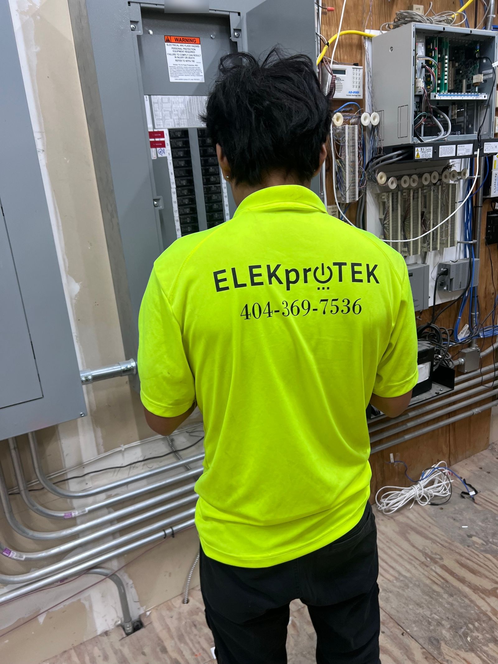 Technician in a lime shirt seen from behind at a large electrical panel, with a low-voltage wiring rack and conduit runs beside it.