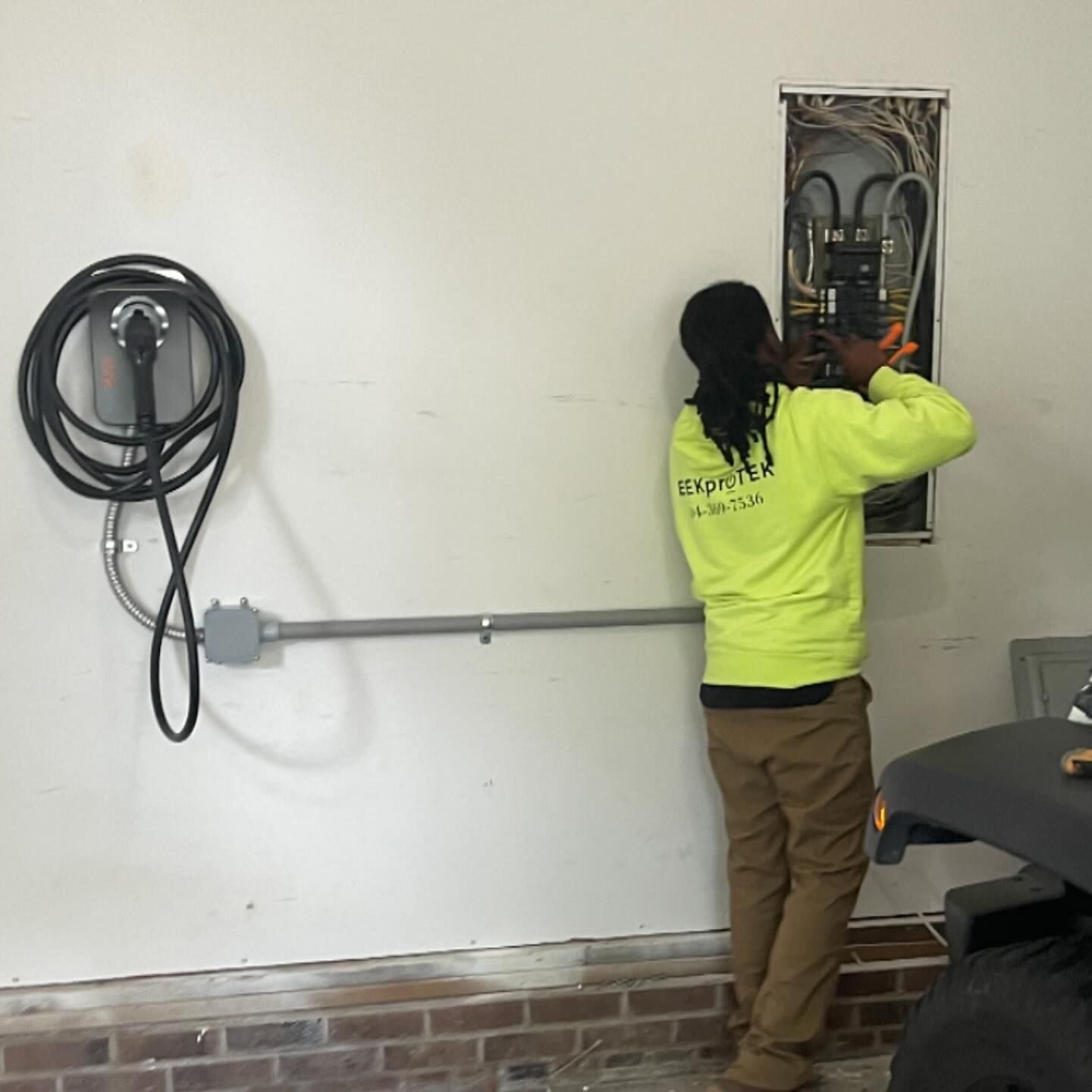 Technician at an open panel in a garage beside a wall-mounted charging unit and a metal conduit run