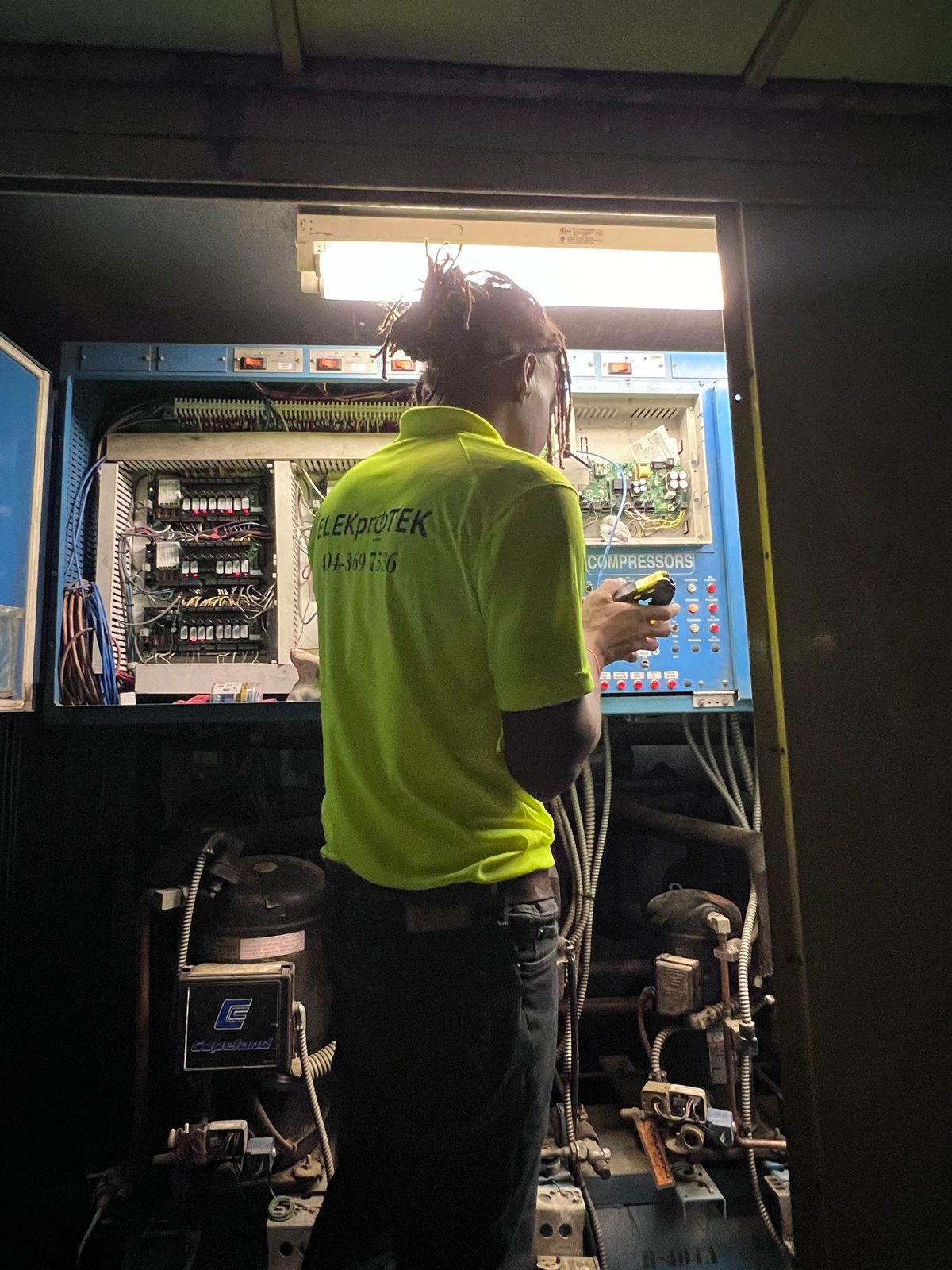 Technician holding a handheld device in front of an open refrigeration control panel above two compressors