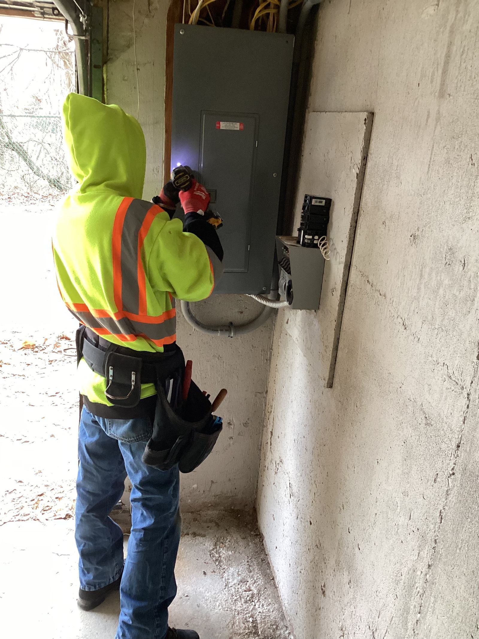 Technician in a hooded lime jacket with orange stripes working at a gray panel on a concrete wall.