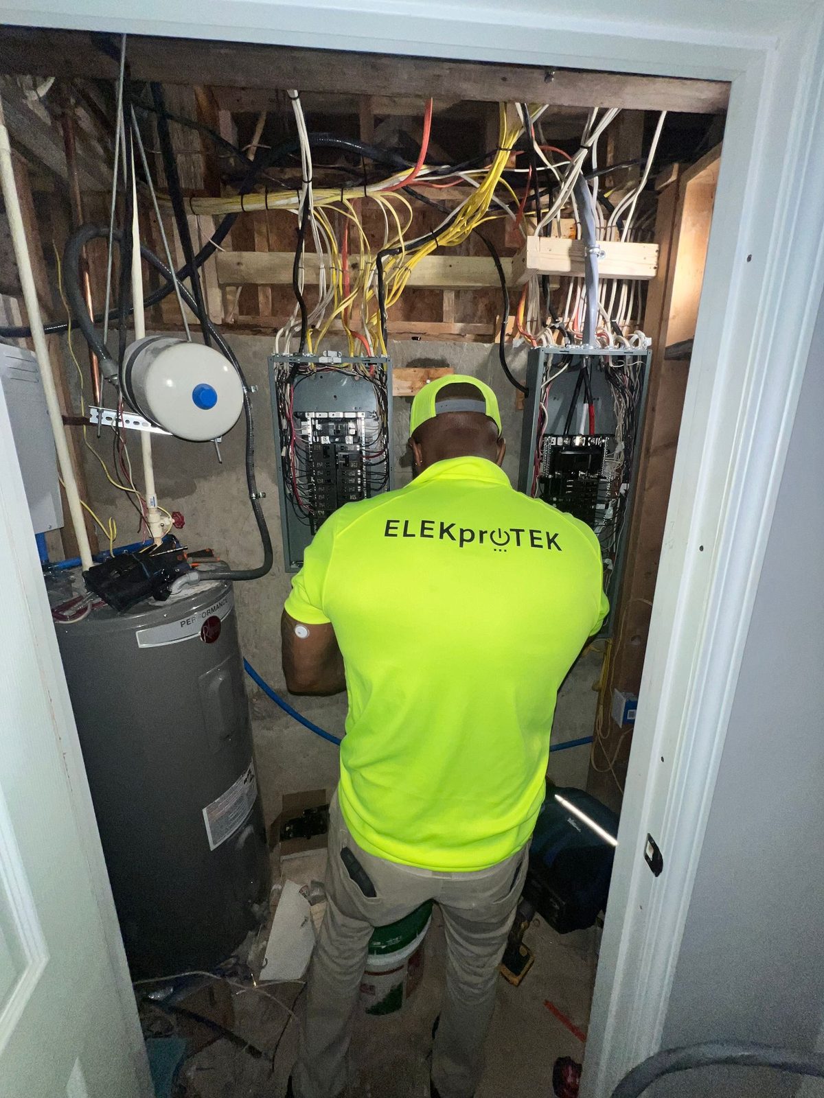 Technician in a lime shirt working in a narrow electrical closet with two panels and a water heater