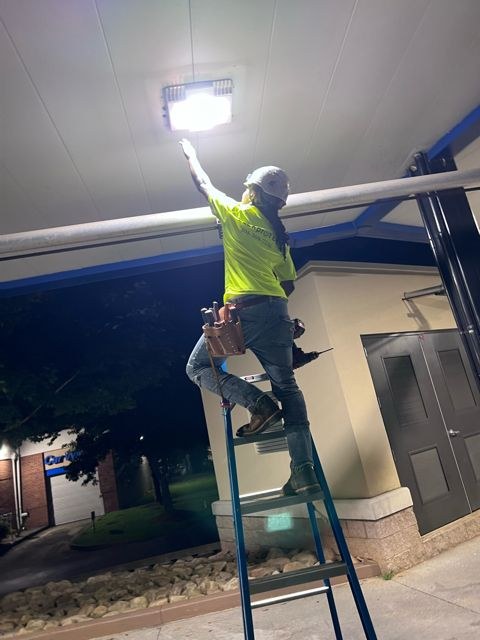 Technician on a ladder reaching up to a bright LED fixture under a canopy at night.