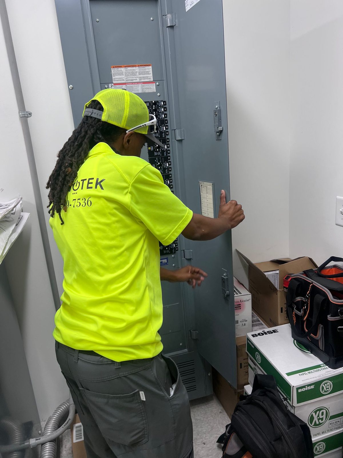 Technician in a lime safety shirt opening the door of a tall panelboard in a small electrical room