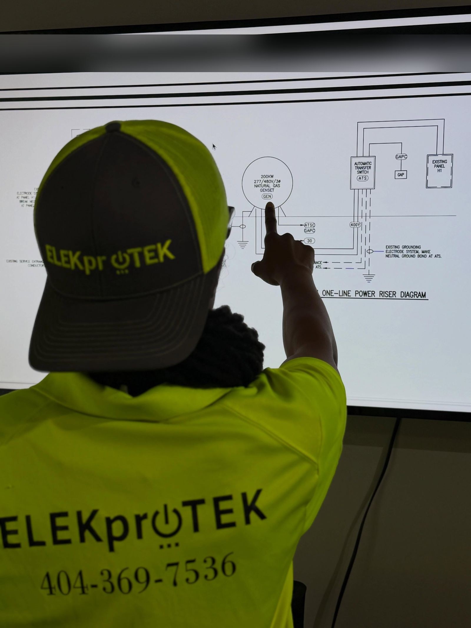 Technician in a cap pointing at a one-line power riser diagram on a large screen.