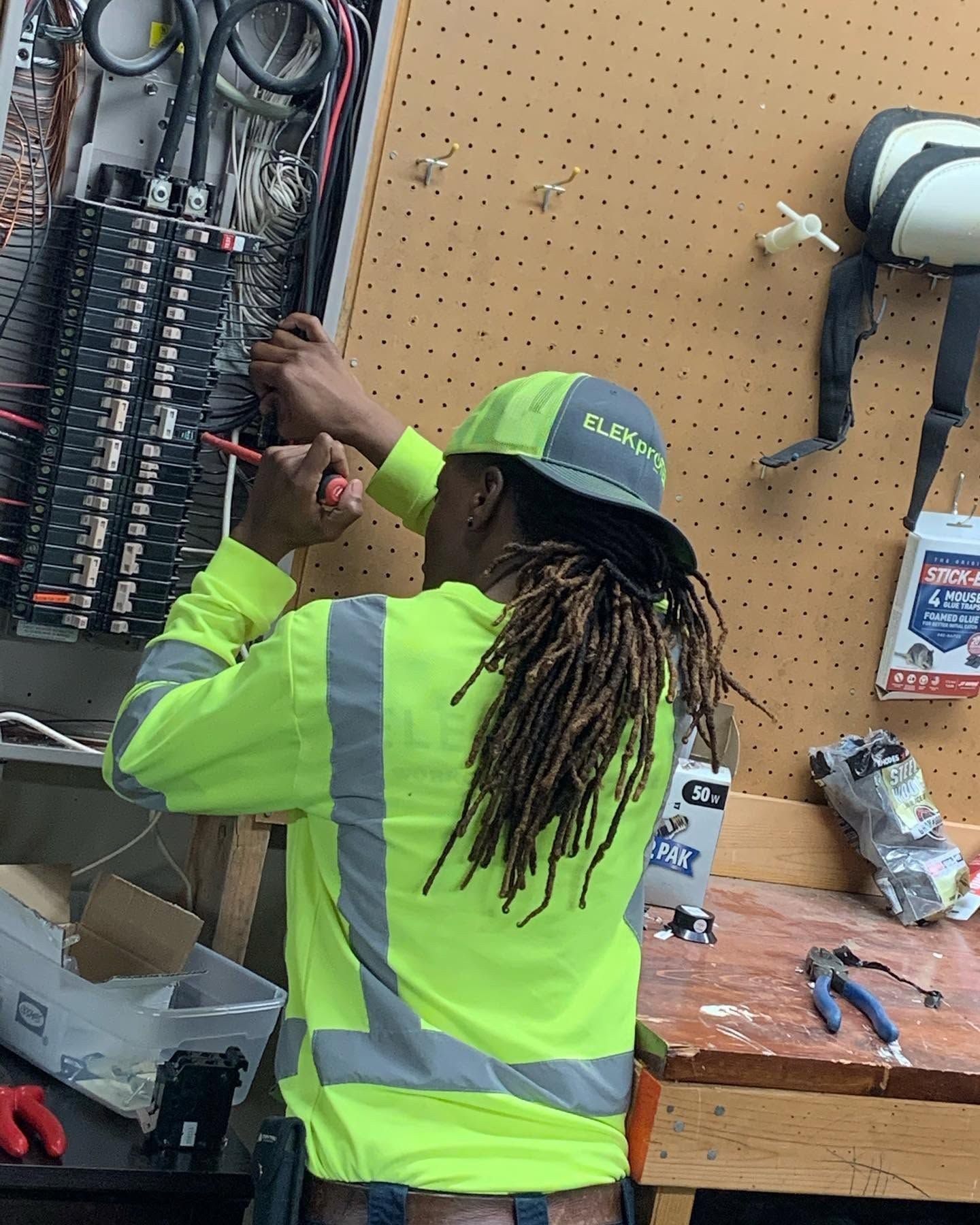 Technician in a reflective jacket terminating a wire in a breaker panel mounted on a pegboard wall.