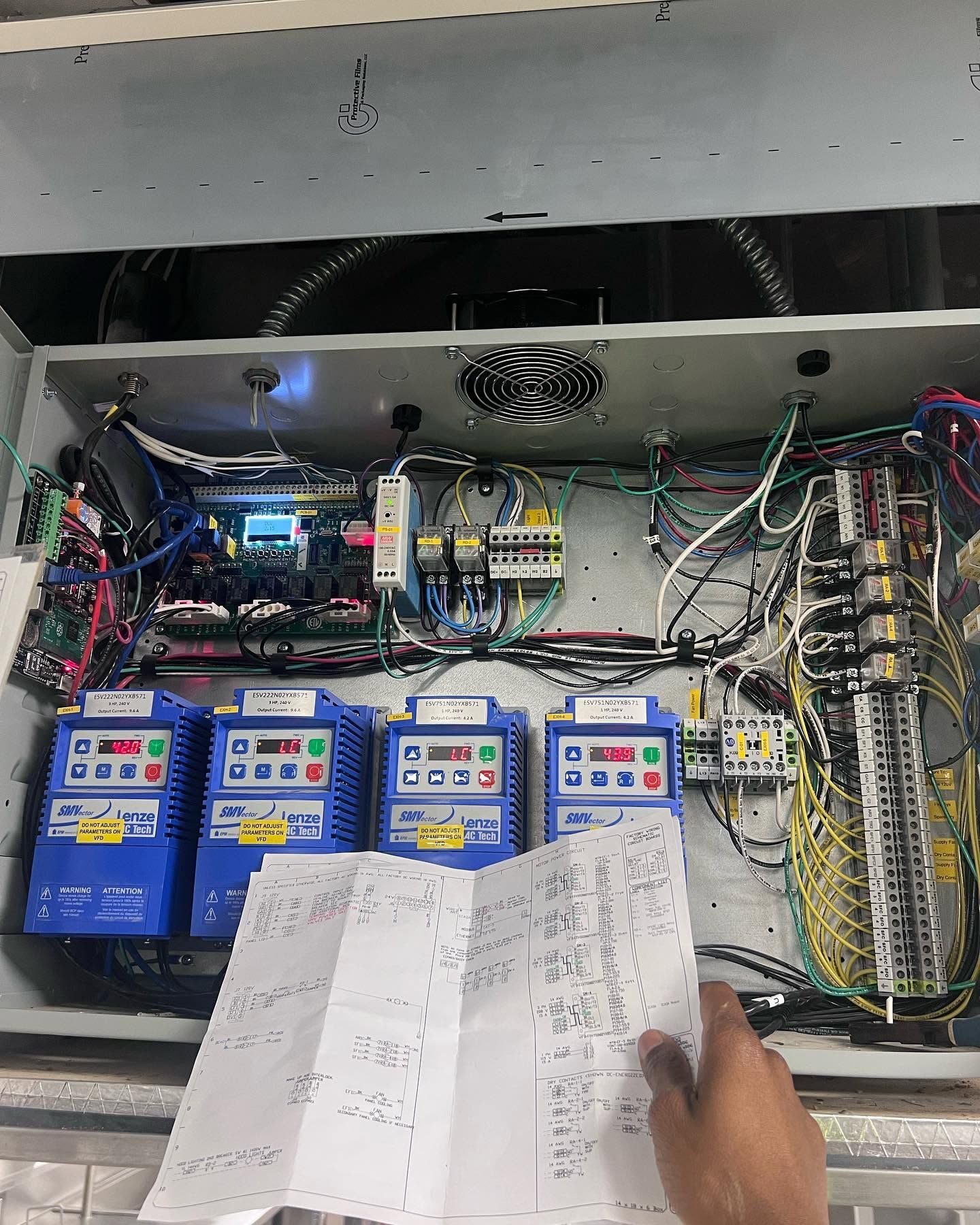 Open control enclosure with four blue variable frequency drives, terminal strips and a hand holding a wiring schematic