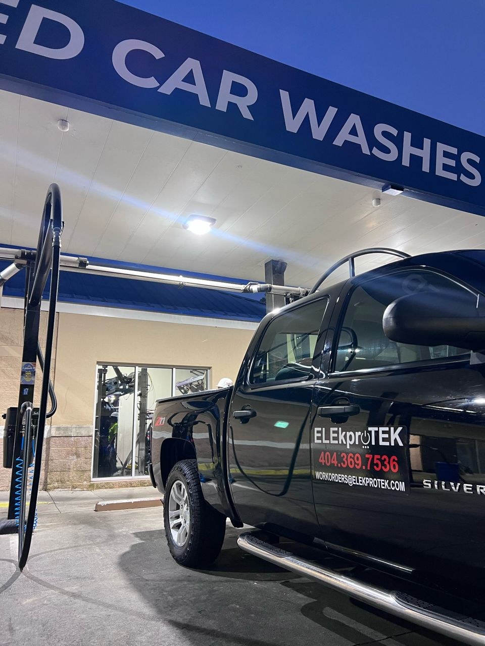 Black work truck parked under a lit commercial canopy at dusk, with a bright LED fixture on the ceiling above.